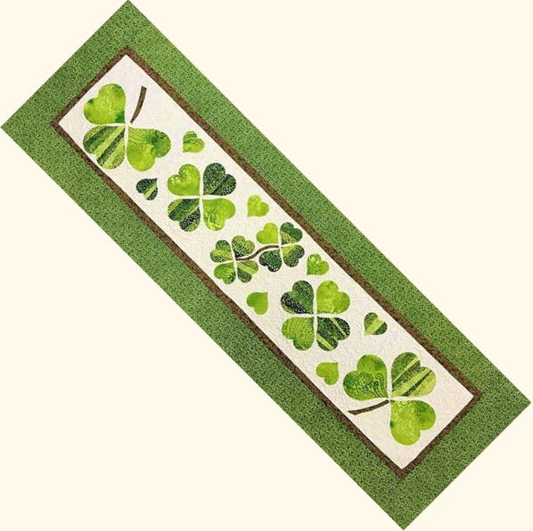 Lucky in Love Table Runner A rectangular, green-bordered quilted St Patrick’s Day table runner pattern featuring appliquéd four-leaf clovers and small green hearts on a white background, displayed at a diagonal angle.