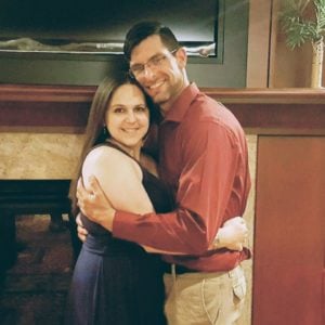 A couple smiling and hugging each other while standing indoors in front of a fireplace. The woman is wearing a dark dress, and the man is wearing a red shirt and khaki pants.