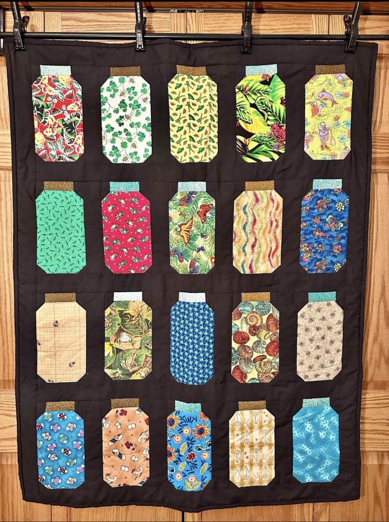 A quilt with 20 Jar Quilt Block shapes, each filled with different colorful fabric patterns, arranged in four rows of five on a black background. The quilt is hanging in front of a wooden wall.