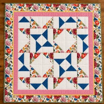 May-Day-Quilt A colorful quilt with a floral border, pink inner border, and Flying Goose Quilt Block patterns in blue, white, and floral prints arranged in squares and triangles, laid on a wooden floor.