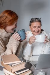 A warm, friendly image of two women enjoying coffee and chatting while using a laptop, surrounded by books, showcasing social interaction and digital connectivity.