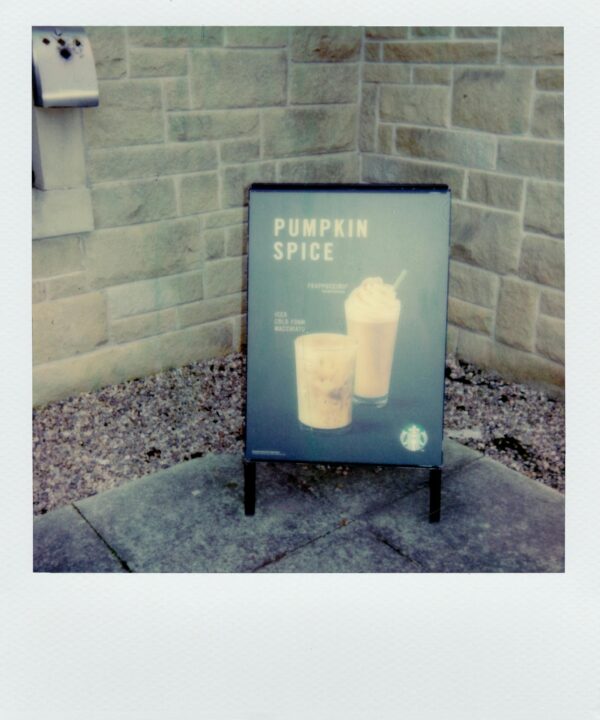 Polaroid photo of a coffee shop's pumpkin spice sign against a stone wall. Lisa from Pexels.