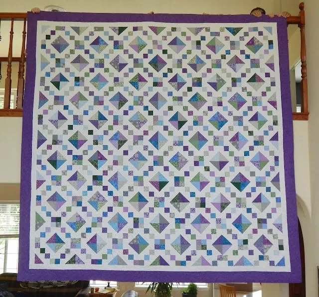 Colorful patchwork quilt with intricate geometric patterns and purple border hanging in a cozy home interior.