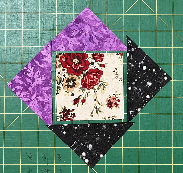 Colorful fabric quilt block pieces with floral, galaxy, and purple patterns on a green cutting mat for quilting and scrapbooking projects.