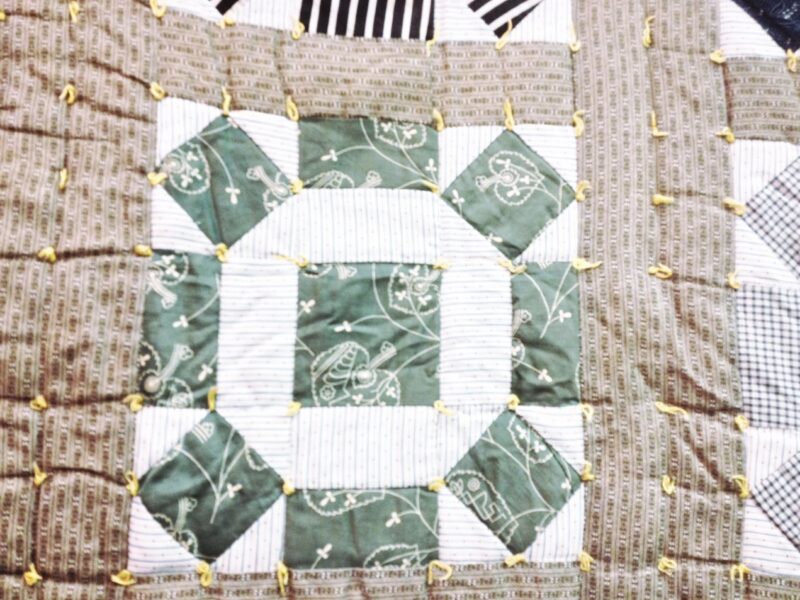 A close-up of an antique quilt, c1900, featuring green, white, and beige patterned fabric squares with yellow yarn ties. The patchwork design includes geometric shapes, floral prints, and a hint of the Wagon Wheel Quilt Block style.