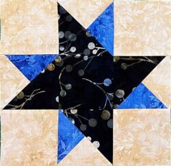 A Quilt Block with 3-part QST features a central eight-pointed star in black fabric with a circular pattern, accented by blue triangles and beige corner squares, creating a striking geometric, patchwork effect.