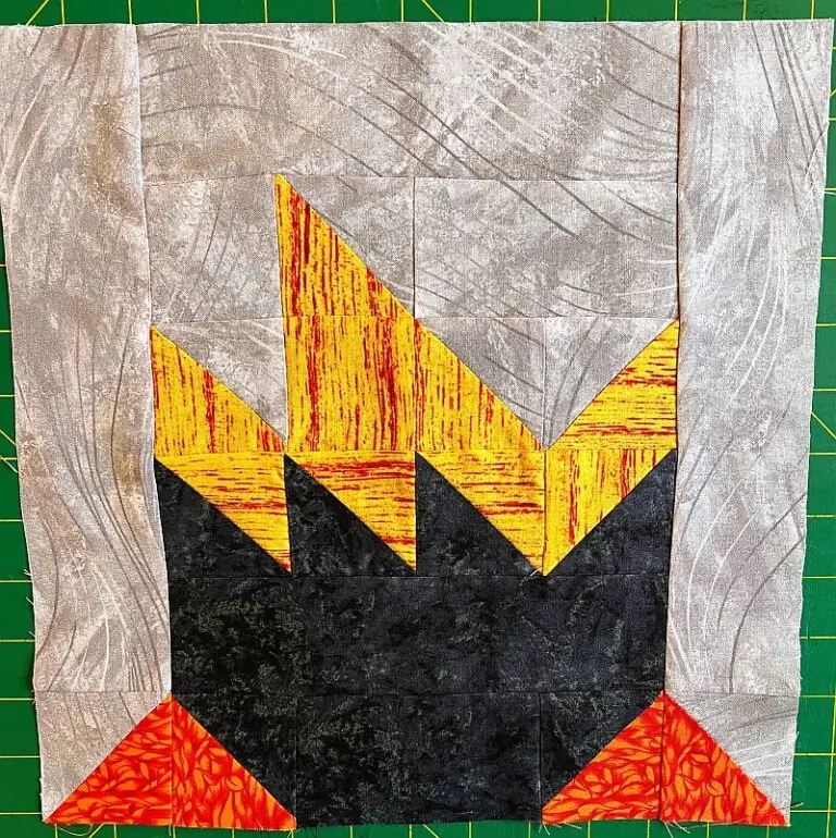 A quilt block features an cauldron design with gray fabric at the top, jagged yellow and orange shapes resembling flames, black fabric below, and red triangles at the bottom corners, all arranged in a 12" square block.