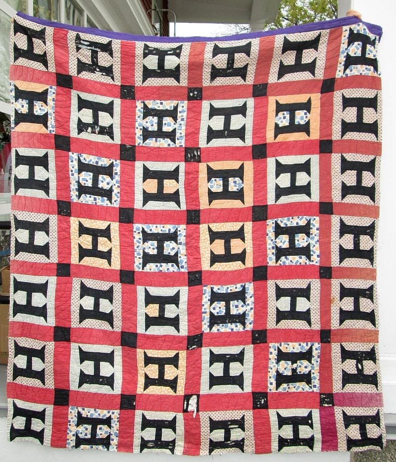 A large, handmade quilt with a geometric pattern—the Hello! Mystery Quilt Introduction—features black, red, blue, and white fabrics in repeating squares and H-shaped designs, hanging outdoors in front of a white porch.