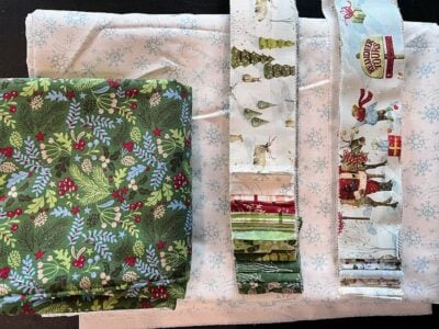 Stacks and strips of holiday-themed fabric in various patterns, including green foliage with berries, Christmas trees, and festive scenes, are arranged on a white background with blue snowflakes.