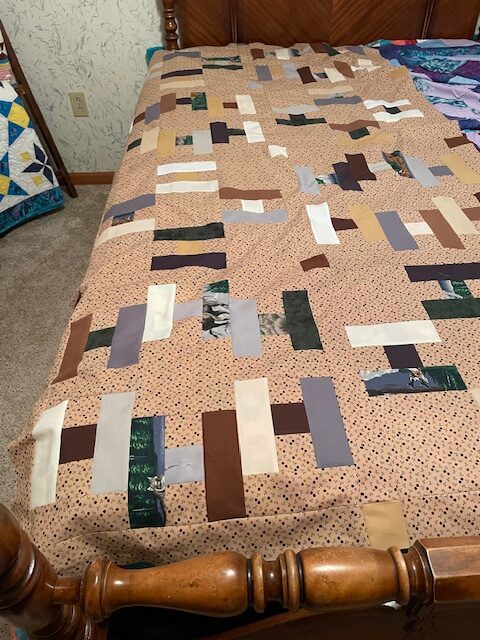 CarolG Hello! This Mystery Quilt Picture Page features a patchwork quilt with rectangular fabric pieces in tan, brown, cream, purple, and green laid on a wooden bed in a cozy, carpeted bedroom. The quilt has a random, modern design.