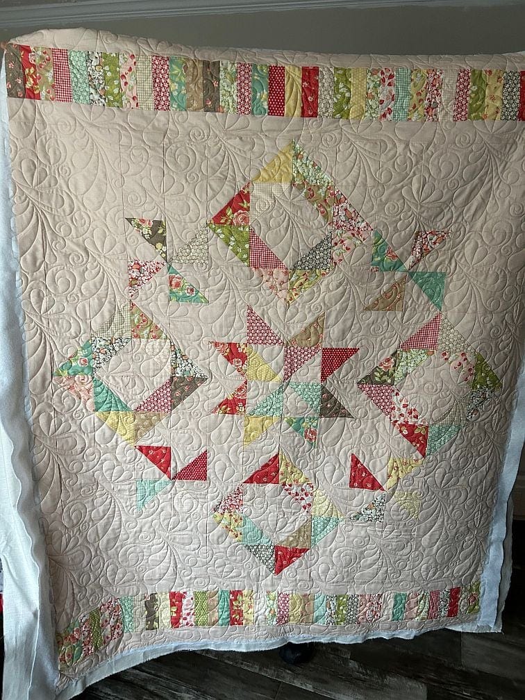 DITS- Marylyn A handmade quilt featuring colorful patchwork geometric patterns and a border of striped fabric, displayed upright. The quilt has intricate stitching with swirls and curved designs on a white background.