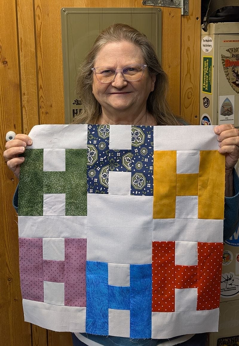 Hello-Debbie-H-block A smiling person with glasses holds up a quilt block featuring six large H-shaped patterns in different colors and prints, including green, blue, yellow, purple, light blue, and red, against a wooden background.