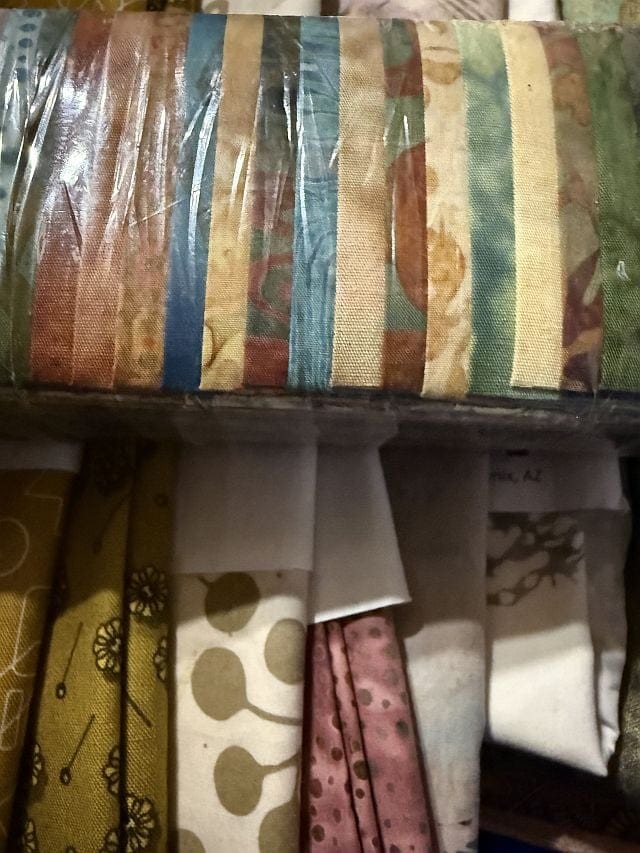 Hello-Diane-C A stack of colorful, patterned fabric strips wrapped in plastic sits above another row of folded fabrics in earthy tones and floral or geometric prints.