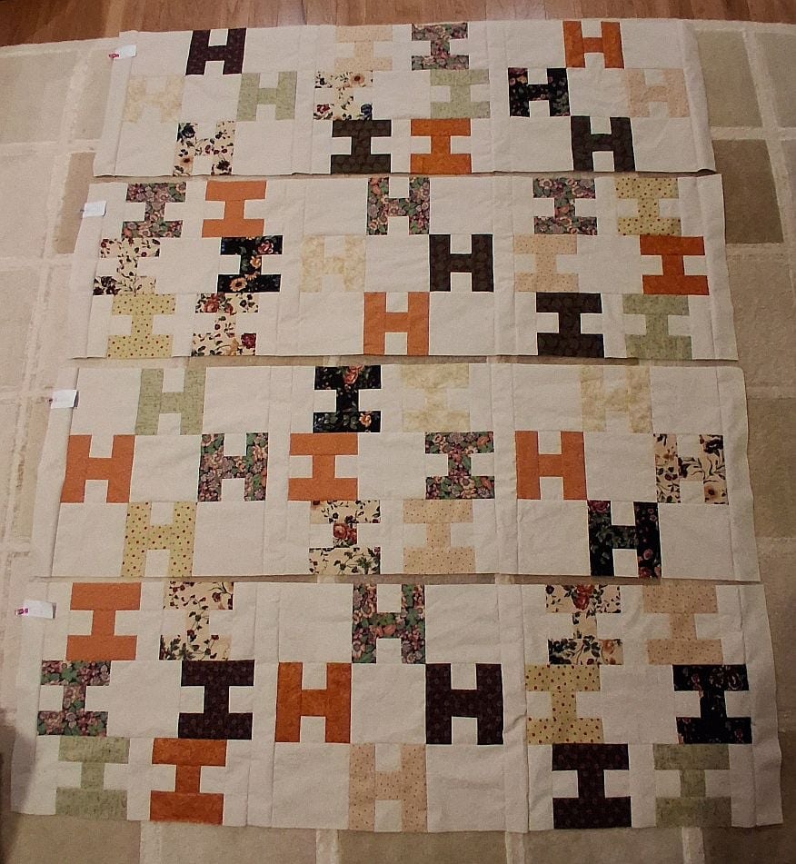 Hello-Gayle-R-blocks A quilt laid out on a beige surface, featuring rows of blocks in an H pattern with various colors and prints, including orange, brown, cream, yellow, and floral fabrics.