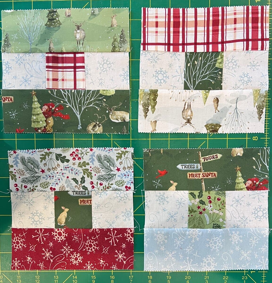 Four quilt blocks made from festive fabric with winter and holiday patterns, including trees, deer, rabbits, snowflakes, berries, and plaid, arranged on a green cutting mat with yellow grid lines.