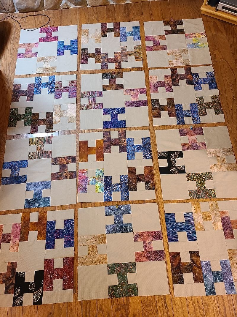 Hello-JanetH-top Several quilt blocks with assorted colorful patterns and neutral backgrounds are laid out on a wooden floor in a grid-like arrangement, ready to be sewn together into a quilt top.