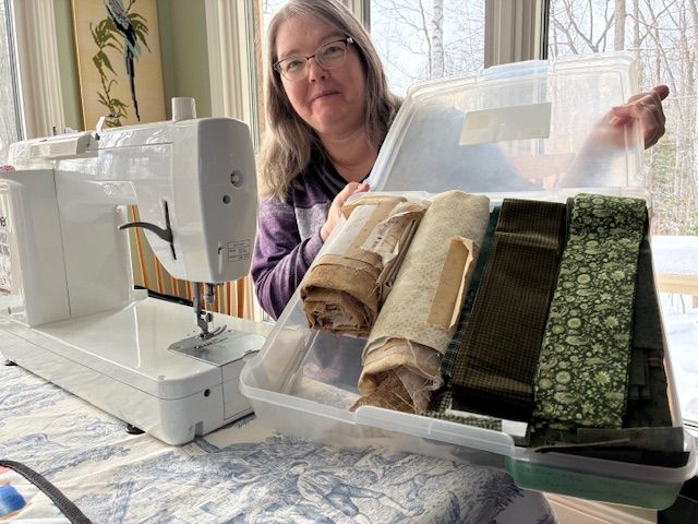 A woman sits at a sewing machine by a window, holding up a clear plastic bin filled with neatly folded fabric in various shades of green and beige. Snow and trees are visible outside.