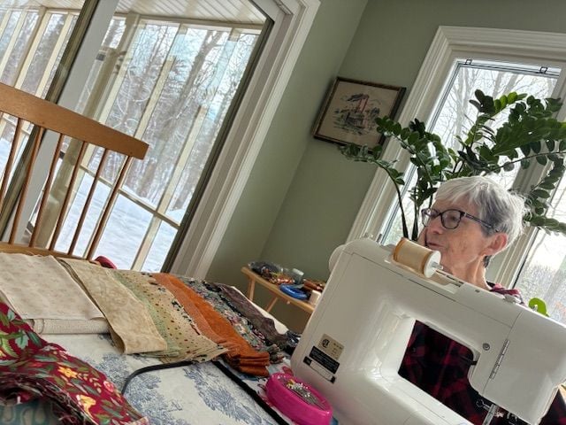 An older woman with short gray hair is sewing at a table using a white sewing machine. She is surrounded by colorful fabric strips and sits by large windows with a snowy outdoor view.