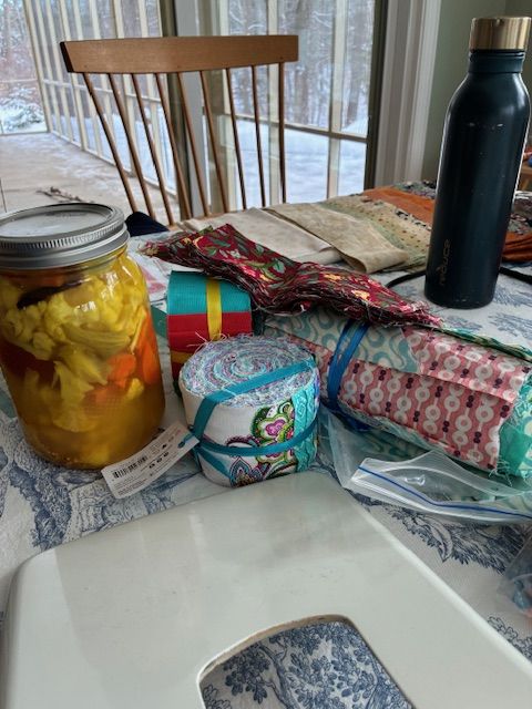 A table holds a jar of pickled vegetables, rolls of colorful fabric, a water bottle, sewing supplies, and a white electronic device. Sunlight streams through large windows in the background, revealing a snowy landscape outside.