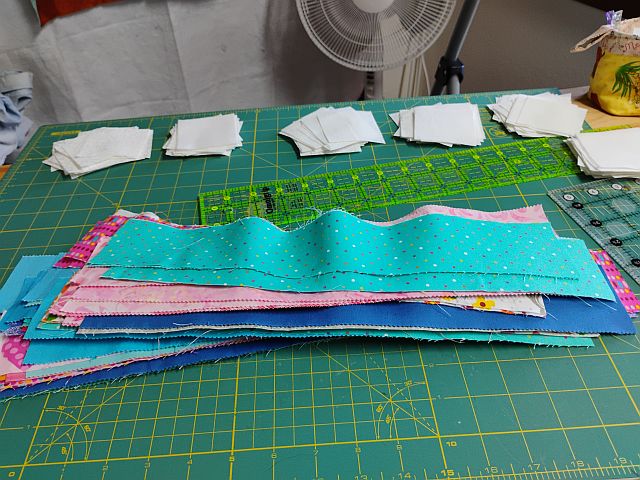Hello-KathrynL-strips A stack of colorful fabric strips and squares lies on a green cutting mat, with neatly arranged white fabric squares and quilting rulers in the background. A fan and a potted plant are also visible.