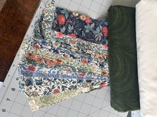 Hello-KathyO-fabrics A patchwork piece of fabric with various floral patterns lies on a cutting mat next to a folded piece of dark green fabric and a pale cream fabric. A ruler is visible along the edge of the mat.