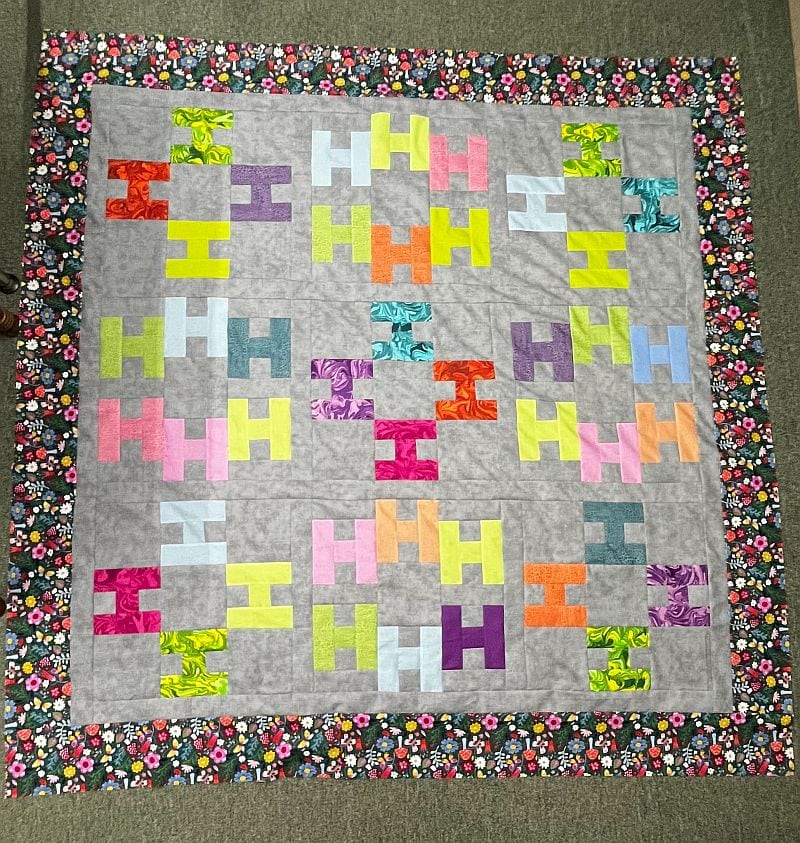 Hello-KellyM-finished A colorful quilt with an H pattern in various bright colors on a light gray background, bordered by a floral fabric with multicolored flowers, displayed on a green carpet.