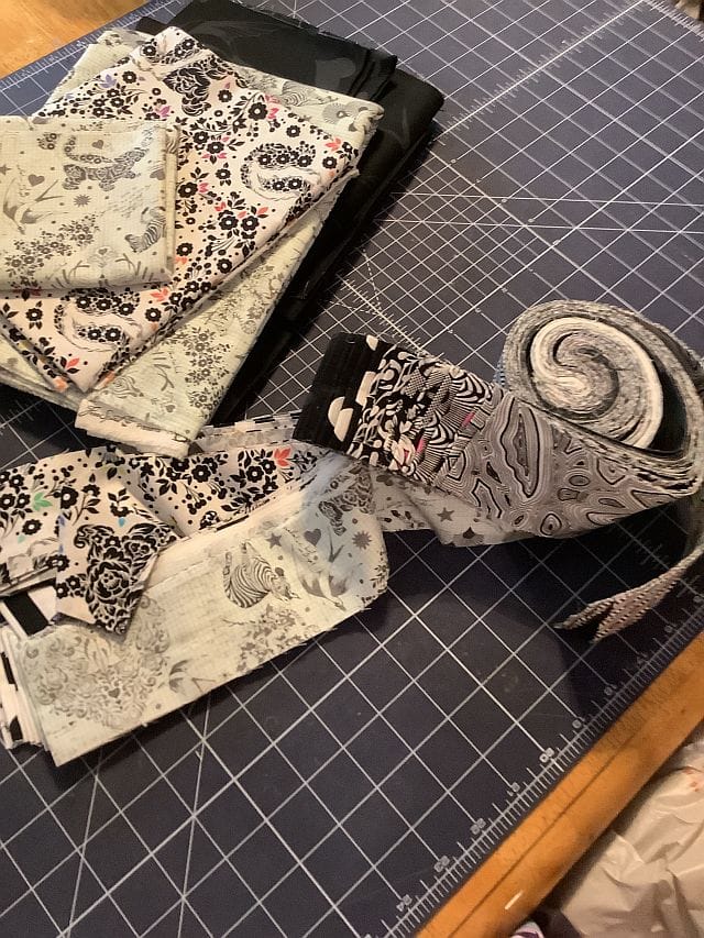 Hello-Margaret-M-fabric Quilting or sewing fabrics in various black, white, and gray patterns are laid out and rolled on a cutting mat with a grid and ruler markings, atop a wooden table.