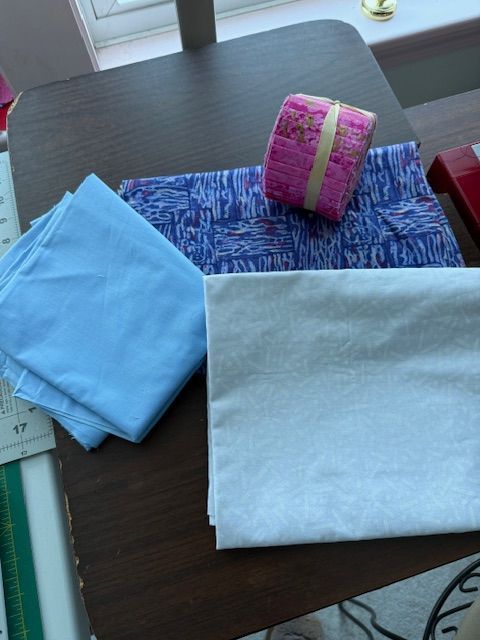 Hello-MarianC-fabrics A table with folded light blue and white fabrics, a folded blue patterned fabric, and a pink floral ribbon roll placed near a window.