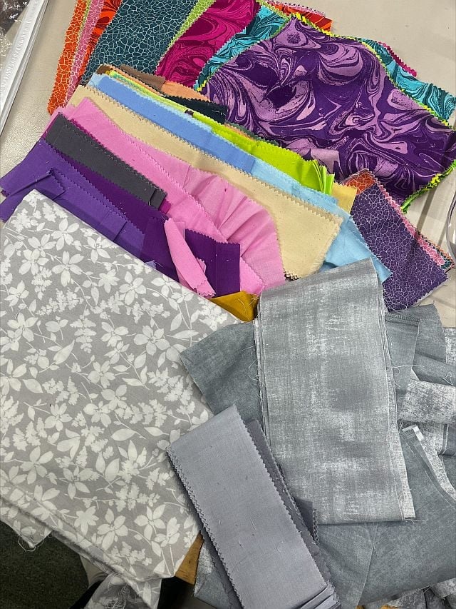 Hello-Marion-fabrics A variety of colorful fabric swatches, including patterned, solid, and textured pieces, are spread out on a table. The fabrics feature shades of purple, blue, pink, yellow, white, and gray.