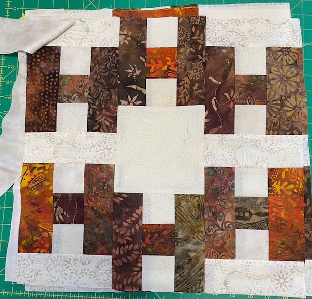 Hello-Mimi-H-block A patchwork quilt block featuring earthy batik fabrics in brown, orange, green, and cream. The design has rectangular strips and a large, solid cream square in the center, displayed on a green cutting mat.