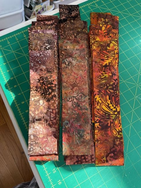 A stack of colorful, patterned fabric strips with batik designs lies on a green cutting mat, which has a yellow grid and ruler markings. Sunlight shines on the fabrics, highlighting their earthy tones.