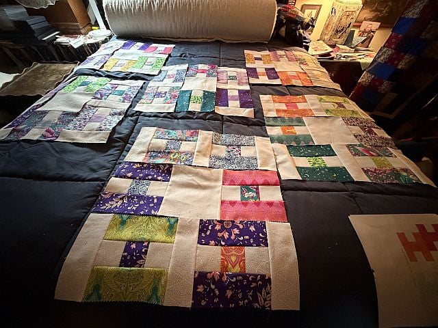 Hello-NancyC-Hs A patchwork quilt with colorful patterned fabric squares is laid out on a dark bedspread in a cozy, cluttered room with books and various items in the background.