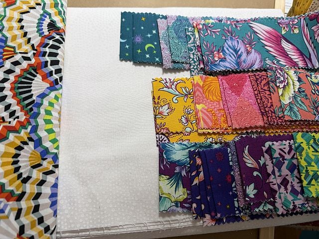 An assortment of colorful fabric swatches with various patterns, including florals, geometric shapes, and celestial motifs, is laid out on a surface next to a white textured fabric.