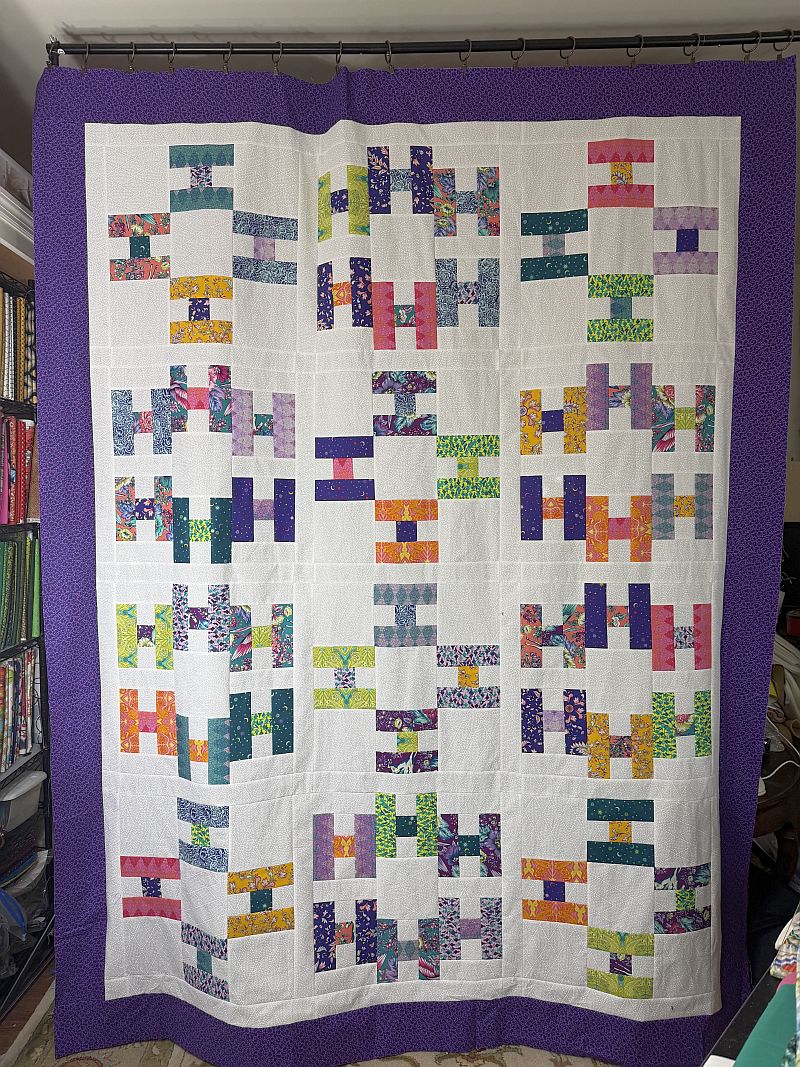 Hello-NancyC-finished A colorful handmade quilt with a white background and purple border, featuring various geometric patterns made from vibrant, multicolored fabrics arranged in rectangular and square shapes.