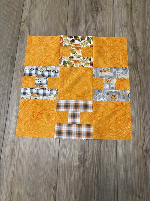 Hello-Pam-H A quilt block with nine squares, featuring mostly orange fabric and accents of patterned fabric with cars, plaid, and autumn leaves, is laid out on a wooden floor.