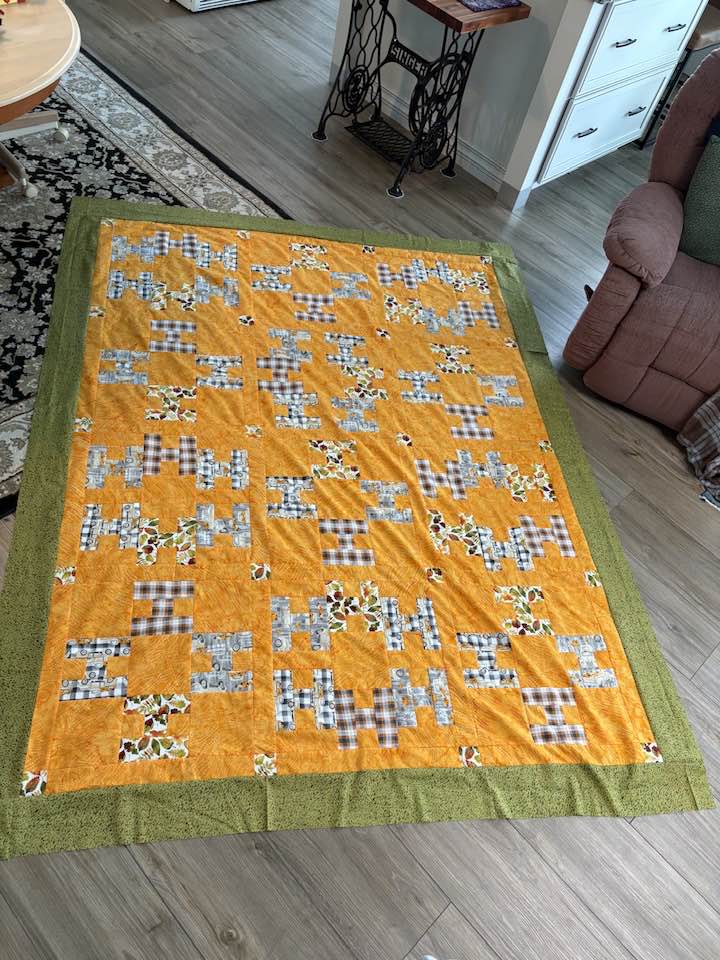 Hello-Pamela-R A handmade quilt with a green border and bright orange center features geometric patterns in gray plaid and floral prints, laid out on a wood floor next to a brown recliner. Perfect for your Hello! Mystery Quilt Picture Page.