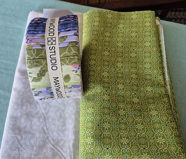 Hello-StephanieD-fabrics Three pieces of fabric: a folded white fabric, a green patterned fabric, and a rolled bundle of floral fabrics labeled Maywood Studio, all arranged on a blue surface.