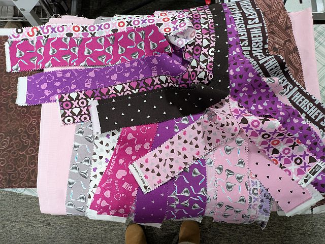 A variety of rectangular fabric swatches in shades of pink, purple, and brown, featuring patterns of hearts, swirls, XOXO text, and Hersheys branding, spread out on a table.