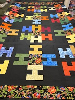 Hello-SueI A colorful quilt, featured on the Hello! Mystery Quilt Picture Page, displays a repeating pattern of “H” shapes in solid and patterned fabrics on a black background, bordered by a vibrant floral print.