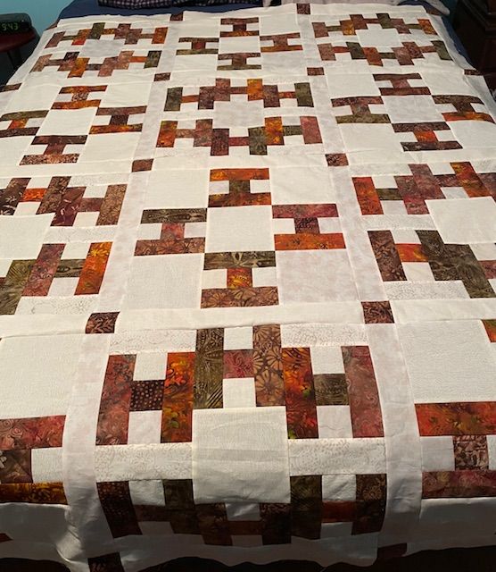 Hello-quilt-top-Mimi-N A handmade quilt with a pattern of rectangular blocks in earthy tones—browns, oranges, and creams—laid out on a bed. The quilt features repeating geometric designs with a patchwork style.