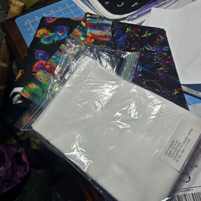 Wanderlust-Beth-D-fabrics A plastic bag containing white fabric rests on top of colorful, music-themed fabric pieces on a cluttered work surface with papers and crafting tools.