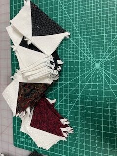 Wanderlust-Carol-Geese Stacks of sewn fabric triangles in black, white, and red sit on a green, gridded cutting mat, arranged in a curving line across the mat.