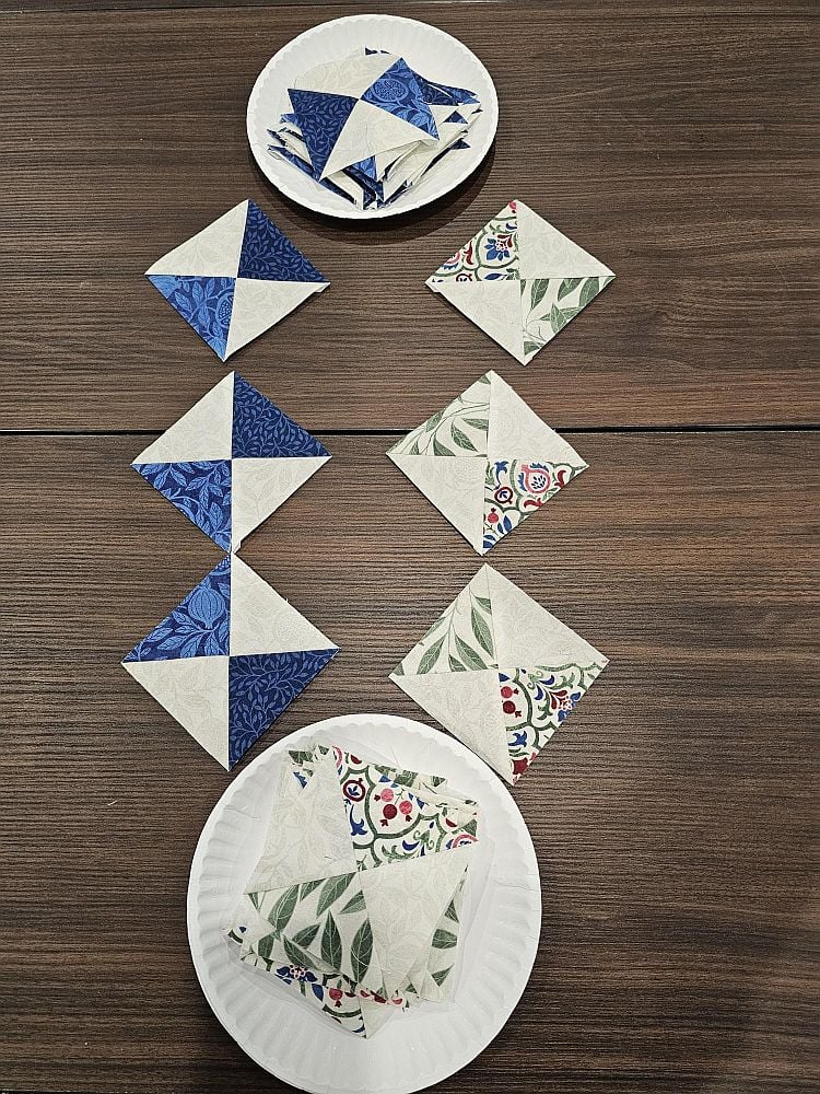 Wanderlust-Daniella-2 Several diamond-shaped quilt pieces in blue, green, and floral patterns are arranged in a vertical line on a wooden table, with two paper plates holding additional quilt pieces at the top and bottom.