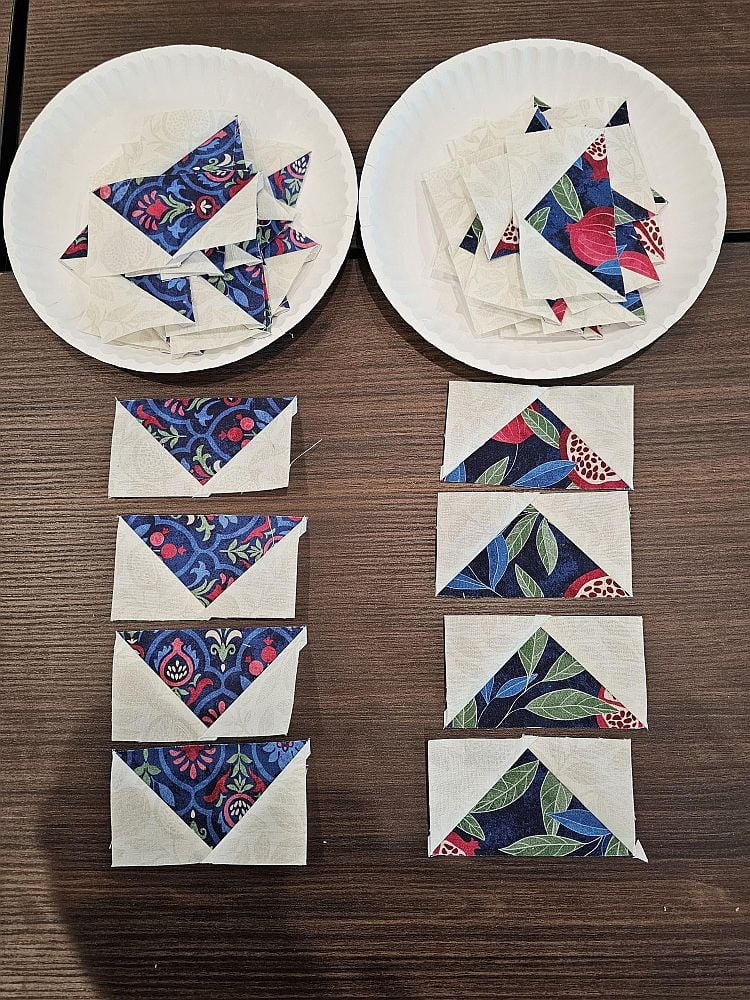 Wanderlust-Daniella-3 Two paper plates hold stacks of small fabric triangles. Below them, quilt blocks made of colorful patterned triangles and white triangles are neatly arranged in rows on a dark wooden table.