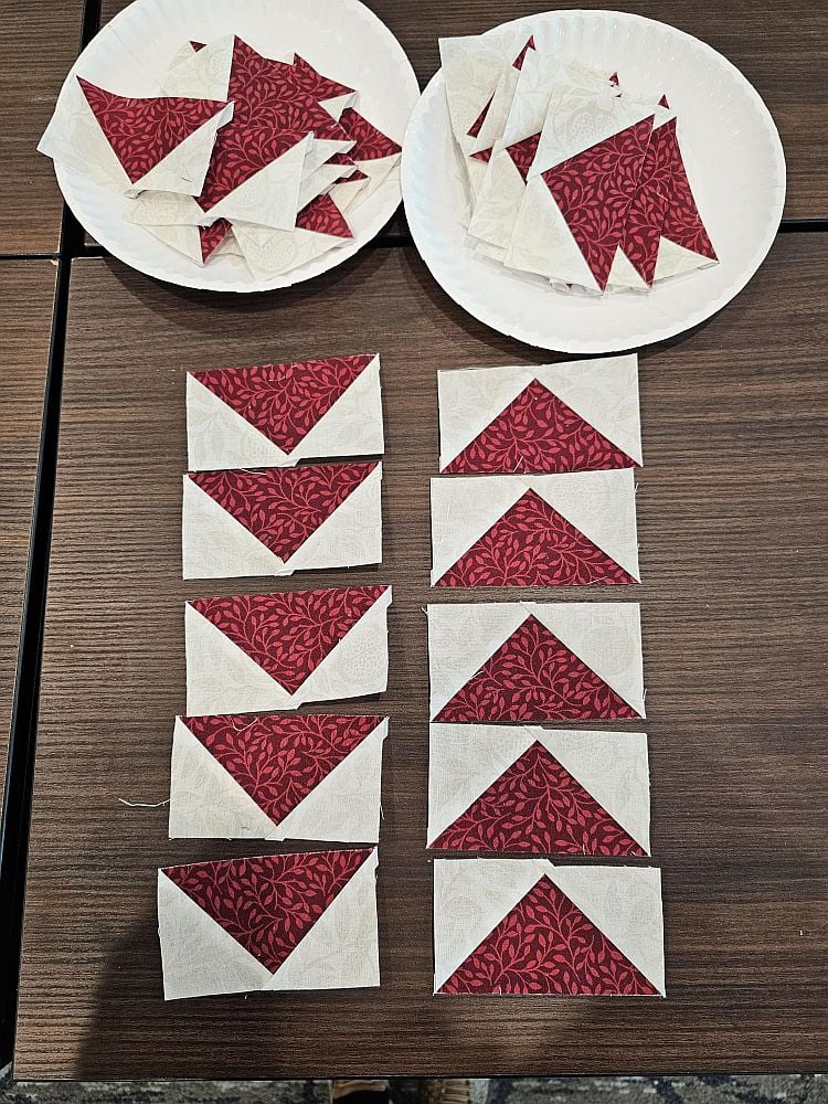 Wanderlust-Daniella-4 Two paper plates hold red and cream triangular quilt fabric pieces. Below them, ten sewn quilt blocks with red triangle centers and cream borders are arranged in two columns on a wooden table.