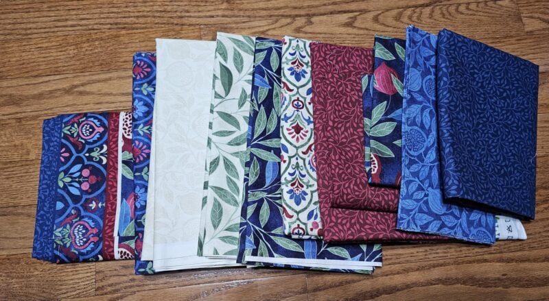 Wanderlust-Daniella-fabrics A set of folded fabric pieces with various patterns, mostly featuring floral and leaf designs in red, blue, green, and cream colors, arranged on a wooden floor.