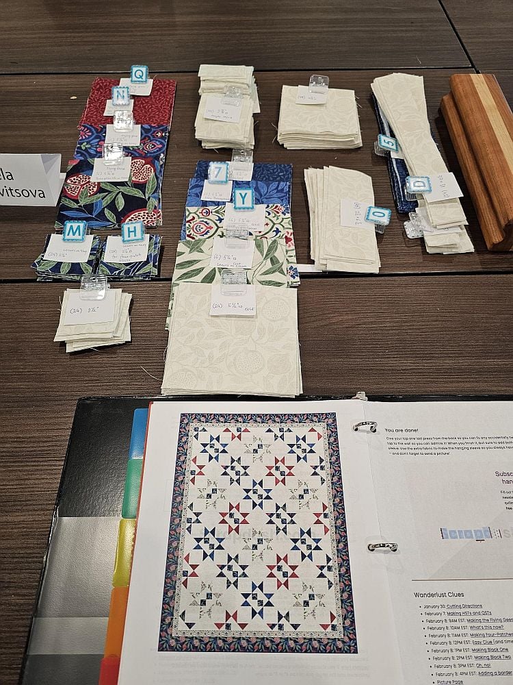 Wanderlust-Daniella-step-one A table with organized stacks of fabric pieces labeled with small cards, next to an open binder showing a quilt pattern with star shapes and instructions.