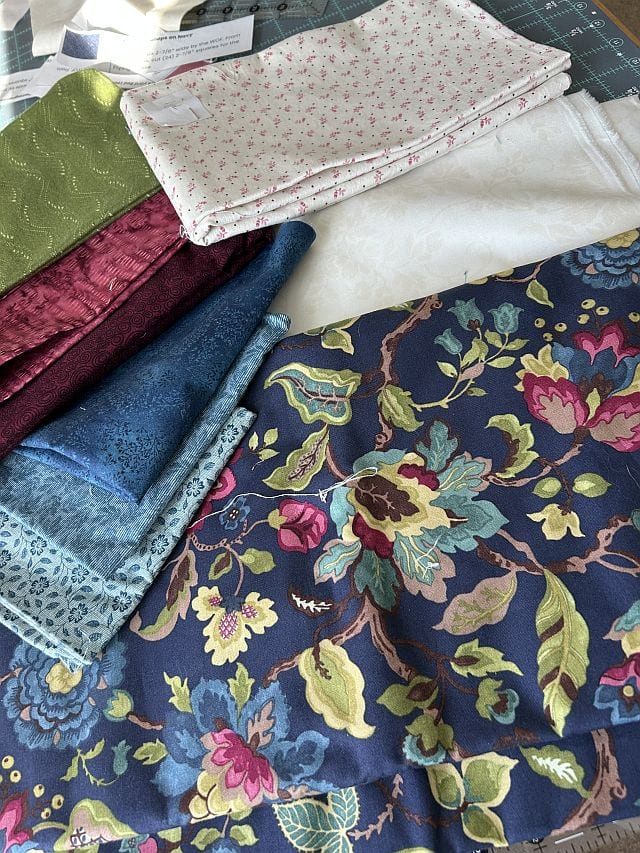 Wanderlust-Fabrics-Marianne-B A variety of colorful fabric pieces are spread out on a surface, including floral, lace, and patterned prints in shades of blue, burgundy, green, white, and pink.