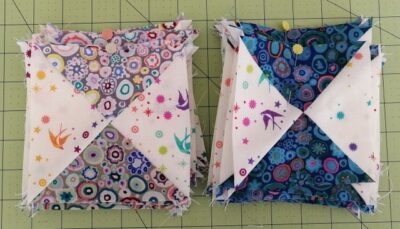 Wanderlust-Kathy-QST Two stacks of colorful quilt blocks with geometric patterns are placed on a green cutting mat. The left stack features lighter fabrics, while the right stack uses darker blue tones with similar prints.