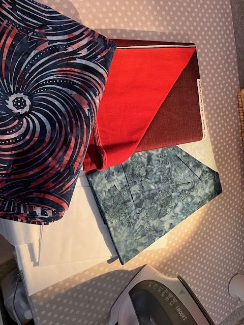 Wanderlust-Nancy-fabrics Several colorful fabric pieces, including red, blue, and marbled patterns, are laid out on a polka-dotted surface near an iron.